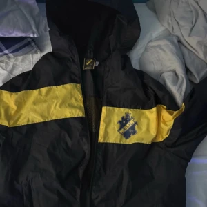 Svart och gul jacka från AIK - Säljer en svart och gul jacka från AIK med dragkedja och huva. Jackan har en gul rand över bröstet och AIK-loggan på framsidan. Perfekt för att visa ditt stöd för laget!