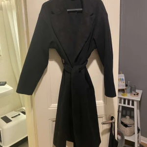 Svart kappa med bälte - Snygg svart kappa med bälte som ger en elegant look. Perfekt för kyligare dagar och passar till många olika outfits. Den har långa ärmar och en klassisk design. Jackan är i använd skick men den är fortfarande fin!