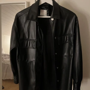 Svart skinnjacka från Bershka - Snygg svart skinnjacka från Bershka med knappar framtill. Jackan har en klassisk krage och långa ärmar. Perfekt för en stilren look.