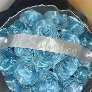 Blå rosor med glitter - En vacker bukett med blå rosor som har en glitter. Rosorna är arrangerade med ett silverfärgat band med texten '18 Sabrina'. Perfekt som en festlig dekoration eller present.