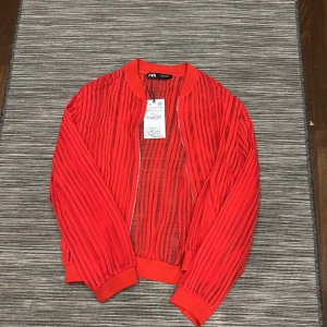 Röd randig bomberjacka från Zara - Snygg röd bomberjacka från Zara med randigt mönster. Jackan har en dragkedja framtill och långa ärmar. Perfekt för att ge din outfit en färgklick! Det är mesh som gör den randiga detaljen. 