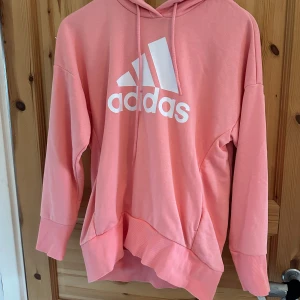 Rosa hoodie från Adidas - Säljer en snygg rosa hoodie från Adidas med det klassiska logotyptrycket i vitt på framsidan. Hoodien har en bekväm passform med långa ärmar och en praktisk huva med dragsko. Perfekt för en avslappnad stil.