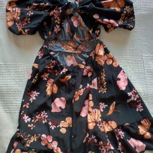 Blommig klänning med öppen rygg - Snygg klänning med blommigt mönster i rosa och orange på en mörk bakgrund. Den har korta ärmar och en öppen rygg med knytning.. Perfekt för en somrig look!