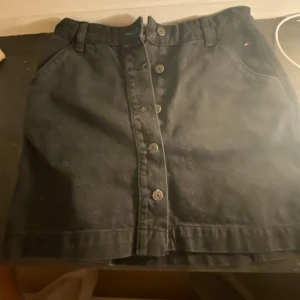 Svart kjol från Tommy Hilfiger - Snygg svart kjol från Tommy Hilfiger med knappar framtill. Perfekt för en stilren look. Kjolen har praktiska fickor och bälteshällor för extra stil och funktionalitet,aldrig andvänt