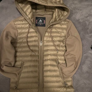 Beige jacka med huva från JOTT - Snygg grön jacka från JOTT med quiltad framsida och huva. Jackan har dragkedja och långa ärmar i ett mjukare material. Perfekt för kyligare dagar. Jackan kanske har en liten udda färg men den är perfekt till ett par ljusa byxor.