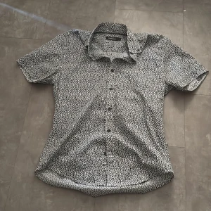 Mönstrad kortärmad skjorta från Dressmann - Säljer en stilren kortärmad skjorta från Dressmann med ett svart och vitt blommigt mönster. Skjortan har knappar framtill och en klassisk krage. Perfekt för en avslappnad men ändå uppklädd look.