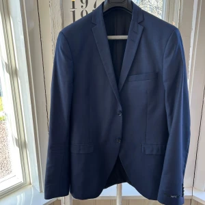 Mörkblå kavaj från Tiger of Sweden - Elegant mörkblå kavaj från Tiger of Sweden. Kavajen har en klassisk design med två knappar framtill och en bröstficka. Byxor ingår med. Kavajen är i storleken 50/50 