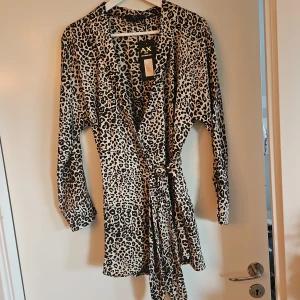 Leopardmönstrad omlott jumpsuit från AX Paris - Snygg leopardmönstrad omlott jumpsuit från AX Paris med långa ärmar och knytband i midjan. Perfekt för att sticka ut med ett djärvt mönster. Klänningen har en v-ringad design som ger en elegant touch.