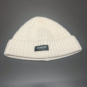 Vit stickad mössa från Dondup - Snygg vit stickad mössa från Dondup, tillverkad i Italien. Den har en ribbad design och en uppvikt kant med en svart etikett framtill. Perfekt för att hålla värmen under kyliga dagar.