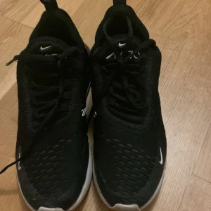 Svarta Nike sneakers - Snygga svarta Nike sneakers med vit sula och diskreta vita detaljer. Skorna har snörning och är perfekta för en sportig look. Modellen är stilren och passar till många olika outfits.