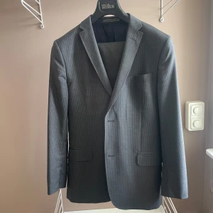Grå randig kostym från J. Lindeberg - Snygg grå randig kostym från J. Lindeberg i slim fit. Kavajen har klassiska slag och byxorna har en stilren design. Perfekt för formella tillfällen.