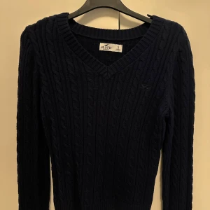 Mörkblå stickad tröja från Hollister - Säljer en snygg mörkblå stickad tröja från Hollister med kabelstickat mönster. Tröjan har en v-ringad hals och långa ärmar, perfekt för kyligare dagar. 