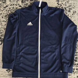 Mörkblå Adidas tröja - Snygg mörkblå träningsjacka från Adidas med vit dragkedja och logga på bröstet. Jackan har långa ärmar och ribbade muddar. Perfekt för sportiga aktiviteter eller som en casual look.