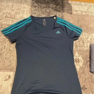 Mörkblå träningstopp från Adidas - Säljer en mörkblå träningstopp från Adidas med korta ärmar och de klassiska tre ränderna i turkos på axlarna. Toppen är tillverkad i ett lätt och andningsbart material, perfekt för träning. Adidas-loggan är tryckt på bröstet. Passar perfekt för en aktiv livsstil!