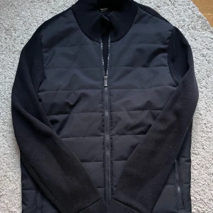 Svart cardigan - Snygg svart quiltad jacka från Massimo Dutti med dragkedja framtill. Jackan har långa ärmar och en stilren design som passar perfekt för kyligare dagar. Perfekt för dig som vill ha en stilren look.