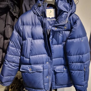Blå dunjacka från Boomerang  - Säljer en klassisk blå dunjacka från Boomerang. Jackan har en pufferdesign med dragkedja och knappar, samt en praktisk huva. Perfekt för kalla dagar med sin varma och isolerande fyllning. Den har flera fickor för extra förvaring.