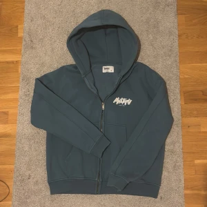 Blå hoodie från Multiply Apparel - Säljer en blå hoodie från Multiply Apparel med tryck både fram och bak. Hoodien har en dragkedja och en stor luva, perfekt för en avslappnad stil. Den är i storlek S och har långa ärmar. Ny pris ligger på 1000kr