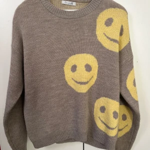 Beige tröja med smileys från Design by Si - Superfin beige tröja med stora gula smileys som ger en glad vibe! Den är stickad och perfekt för höst och vinter. Tröjan har en avslappnad passform och långa ärmar, vilket gör den både bekväm och stilren. Perfekt för att liva upp vardagslooken! Använd fåtal gånger.