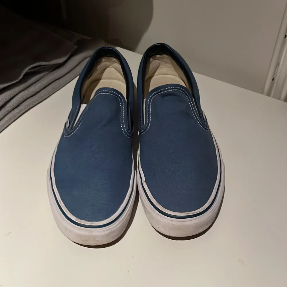 Säljer ett par klassiska blå slip-on sneakers från Vans. De har en vit sula och är i bra skick med lite slitage på sulan. Perfekta för en avslappnad stil och passar till det mesta. Skick 7.5/10. Skor.