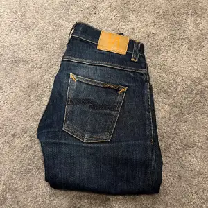 Säljer ett par snygga mörkblå jeans från Nudie Jeans i storlek W30 L32. De har en klassisk femficksdesign med orange sömmar och knappgylf. 