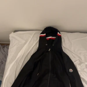 Moncler Hoodie - Säljer en svart Moncler Hoodie med röd och vit detalj i storlek M. Sitter som S! Använd 1 gång. Bättre pris vid snabb affär. 
