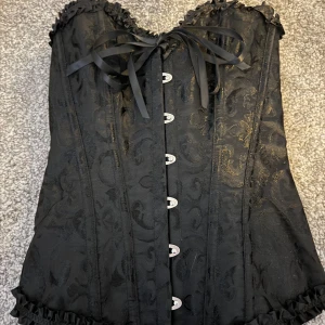 Svart korsett med snörning  - Snygg svart korsett med vackert mönster och detaljer som snörning i ryggen. Den har en elegant volangkant upptill och nedtill samt en söt rosett framtill. Perfekt för festliga tillfällen!