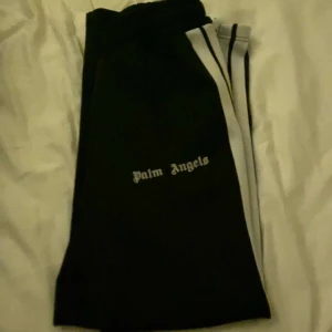 Svarta byxor från Palm Angels - Säljer ett par svarta byxor från Palm Angels med vit text på sidan. De har en sportig look med vita ränder längs benen. Perfekta för en avslappnad stil eller träning. Passar bra till både vardag och fest.