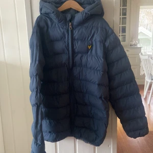 Blå pufferjacka från Lyle & Scott - Säljer en snygg blå pufferjacka från Lyle & Scott i storlek 12/13 år. Jackan har en mysig huva och dragkedja framtill. Perfekt för kalla dagar och håller dig varm och bekväm. Passar både till skolan och fritiden. 🧥💙det enda som är fel på den är ett hål i fickan till högra fickan. Hör av er för fler bilder och priset är inte hugget i sten.