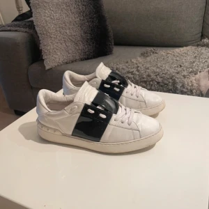 Valentino open/sneakers i vitt och svart - Säljer ett par snygga Valentino sneakers i vitt med en cool svart detalj över snörningen. Skorna är i bra skick och har en stilren design som passar perfekt till både vardag och fest. De är bekväma och har en klassisk look som aldrig går ur tiden.