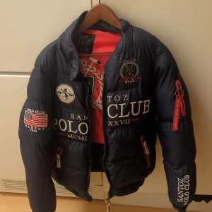 Navy pufferjacka från Santoz Polo Club - Säljer en svart pufferjacka från Santoz Polo Club i okej skick. Jackan har broderade detaljer och dragkedja framtill. Perfekt för höst och vinter! 