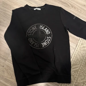 Svart tröja från Stone Island - Säljer en svart tröja från Stone Island i mycket bra skick. Den har ett stort, snyggt Stone Island-tryck på framsidan och en broderad logga på ärmen. Perfekt för höst och vinter, med normal passform och långa ärmar. Grym för både vardag och fest!