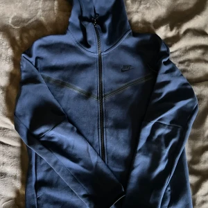 Blå hoodie från Nike - Säljer en snygg blå hoodie från Nike i storlek M. Den har en dragkedja framtill och en praktisk ficka med dragkedja på ärmen. Perfekt för höst och vår, med en stilren design och Nike-logga på bröstet. Passar både till träning och vardag.