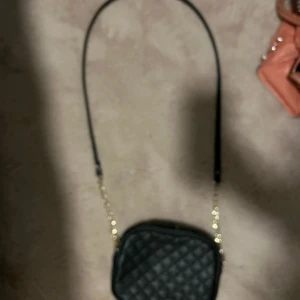 Svart quiltad axelväska - Säljer en snygg svart quiltad axelväska med guldkedja. Väskan är i syntetmaterial och har en praktisk dragkedja upptill. Perfekt för både vardag och fest! 🖤