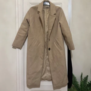 Beige kappa  - Säljer en stilren beige kappa i mycket bra skick. Den har en klassisk design med långa ärmar och enkla knappar framtill. Perfekt för höst och vinter, och funkar lika bra till vardags som till fest. Storlek dam S.