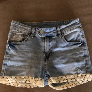 Jeansshorts med spetsdetalj - Säljer ett par snygga jeansshorts i ljusblå denim med fina spetsdetaljer längst ner. De har en klassisk femficksdesign och är perfekta för sommardagar. Shortsen är i bra skick och har en bekväm passform.