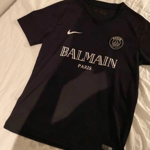 Svart PSG x Balmain fotbollströja - Säljer en svart PSG x Balmain fotbollströja i Dri-FIT material från Nike. Tröjan har korta ärmar och en stilren design med Balmain Paris-logga på bröstet. Perfekt för både träning och vardag! Tvättad endast en gång och är i mycket bra skick.