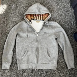 Burberry kofta - Säljer en snygg grå huvtröja från Burberry med klassiskt rutigt foder i huvan. Tröjan har dragkedja och två fickor fram. Perfekt för höst och vår, och den är i mycket bra skick. Passar både till vardags och lite mer avslappnade tillfällen.