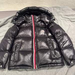 Säljer en snygg svart pufferjacka från Moncler. Den har en cool glansig finish och en röd-vit dragkedja som ger en extra touch. Jackan är perfekt för kalla dagar och har en mysig huva. Inuti finns en rolig serietidningstryck. Perfekt för vintern!