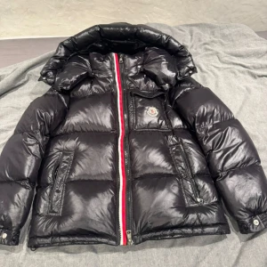 Svart pufferjacka från Moncler - Säljer en snygg svart pufferjacka från Moncler. Den har en cool glansig finish och en röd-vit dragkedja som ger en extra touch. Jackan är perfekt för kalla dagar och har en mysig huva. Inuti finns en rolig serietidningstryck. Perfekt för vintern!
