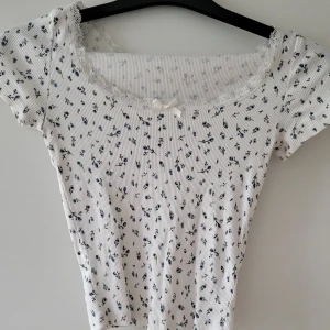 Vit blommig topp från HM - Söt vit topp från Divided med små blå blommor och spetsdetaljer vid halsringningen. Den har en liten rosett framtill och korta ärmar. Storlek: XS  Skickas inslagen i silkespapper i vattentätt paket.   Skicka pm för fler bilder. 