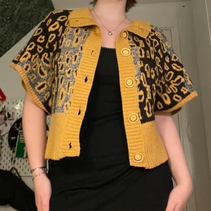 Gul och svart mönstrad kofta - Säljer en unik och färgglad kofta i gult och svart med leopardmönster. Den är stickad och har en ribbad krage samt knappar framtill. Perfekt för att liva upp din höstgarderob! Tillverkad i Turkiet av 70% ull och 30% akryl.