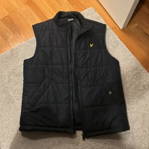 Lyle & Scott väst  - Säljer en snygg svart quiltad väst från Lyle & Scott. Den har en dragkedja framtill och två praktiska fickor. Perfekt för höst och vår när du vill hålla dig varm men ändå se stilren ut. Västen är i bra skick och har en klassisk design med en liten logga på bröstet.