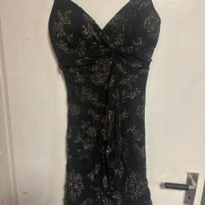 Svart klänning med guldmönster - Säljer en elegant svart klänning med vackert guldmönster. Klänningen har en v-ringad design och är perfekt för festliga tillfällen. Materialet känns lyxigt och faller fint längs kroppen. Perfekt för en kväll ute eller en speciell tillställning!