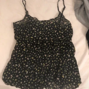 linne - Säljer en söt blommig linne i svart med små vita blommor. Den har smala axelband och en luftig passform, perfekt för sommardagar. Passar både till stranden och en avslappnad dag på stan. 🌸