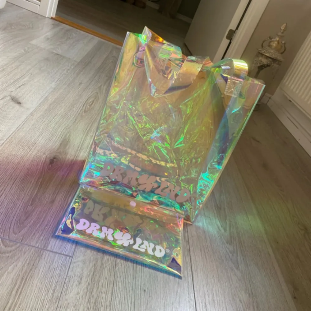 Säljer en supercool holografisk shoppingväska från Drksld. Väskan har en glansig, regnbågsskimrande yta som verkligen sticker ut. Perfekt för att bära med sig allt man behöver och samtidigt se trendig ut. Den har en praktisk knappstängning och är lätt att bära med sig.. Väskor.