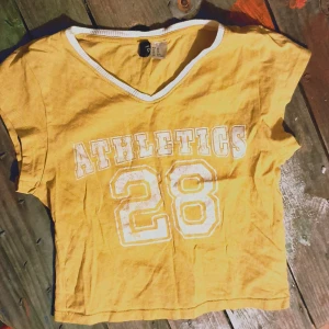 Gul v-ringad t-shirt med tryck - Säljer en cool gul t-shirt från h&m med v-ringning och trycket 'ATHLETICS 28' i vitt. Perfekt för en avslappnad stil eller sportiga dagar. T-shirten är croppad och har korta ärmar. Skön och lätt att bära!