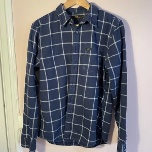 Blå rutig skjorta från Lee - Säljer en snygg blå rutig skjorta från Lee i storlek M. Skjortan har långa ärmar och en klassisk krage. Den är perfekt för både vardag och festliga tillfällen. Materialet känns mjukt och bekvämt, och den har en bröstficka med Lee-loggan. Passformen är regular fit.