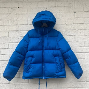 Blå pufferjacka med huva - Säljer en supermysig blå pufferjacka med huva. Perfekt för kalla dagar med sin tjocka vaddering och långa ärmar. Jackan har en dragkedja framtill och dragsko vid huvan för extra skydd mot vinden. Den är i bra skick och redo för vintern!