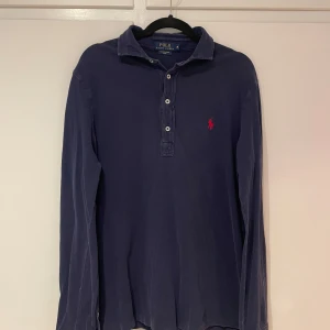 Blå långärmad pikétröja från Ralph Lauren - Säljer en klassisk blå långärmad pikétröja från Ralph Lauren. Tröjan har en krage med knappar och det ikoniska röda broderade logotypen på bröstet. Perfekt för en stilren och avslappnad look.