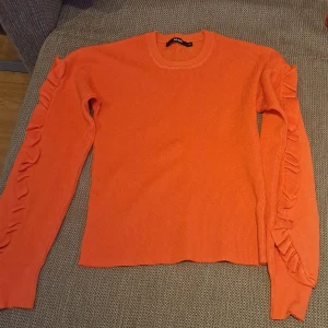 Orange stickad tröja med volangdetaljer - Säljer en snygg orange stickad tröja med volangdetaljer längs ärmarna. Tröjan har en rund halsringning och långa ärmar, perfekt för att liva upp garderoben med lite färg.